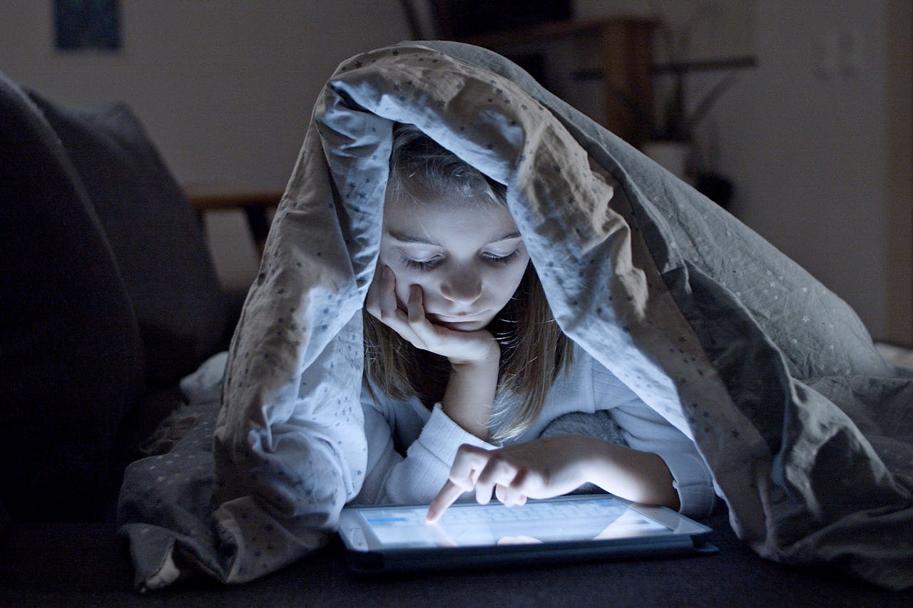 A child using a tablet under a blanket in a dark room, creating a cozy bedtime atmosphere.